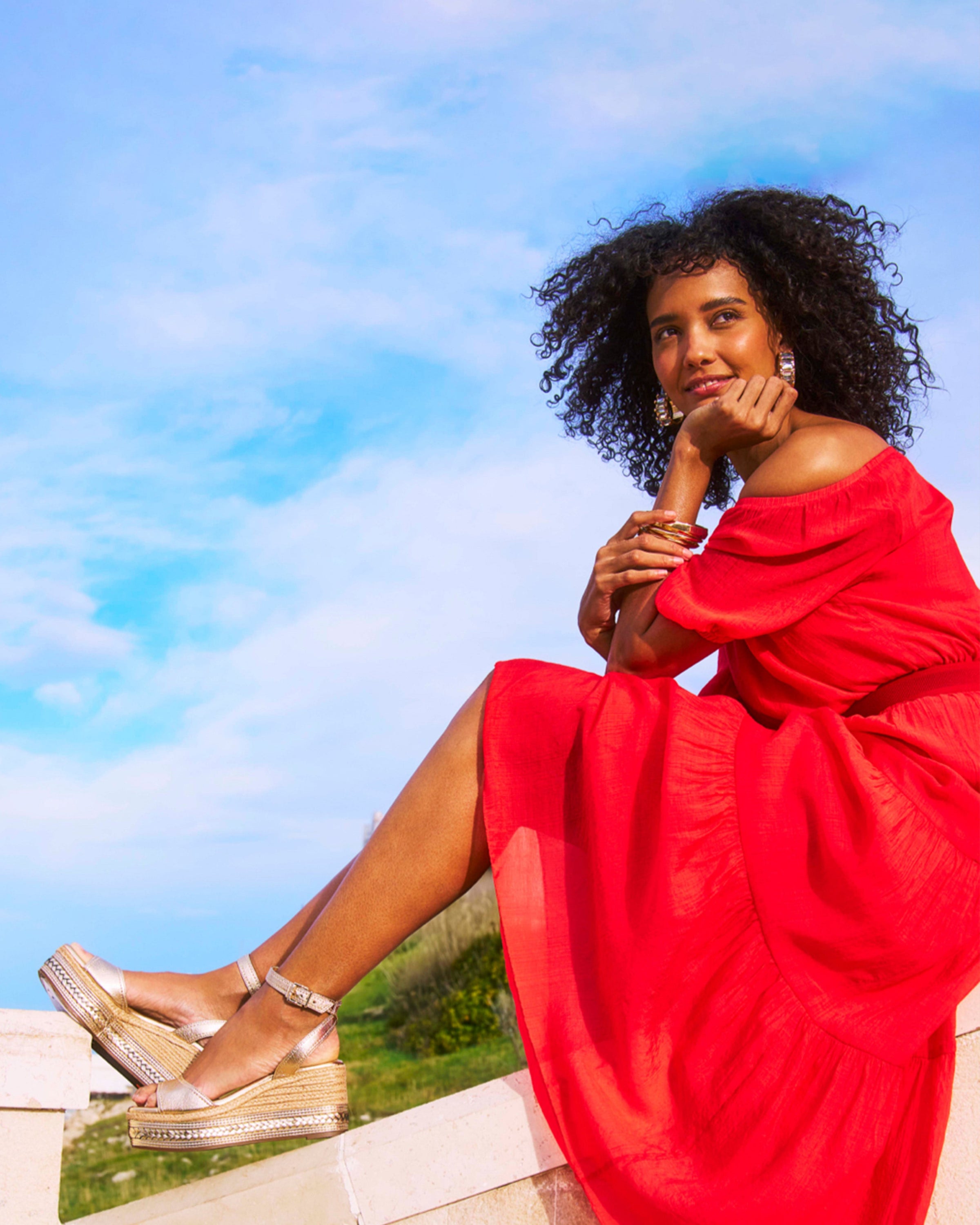 A woman in a bright red dress sits on a stone ledge outdoors, smiling thoughtfully. She has curly hair and wears gold platform sandals. Blue sky and greenery are visible in the background.