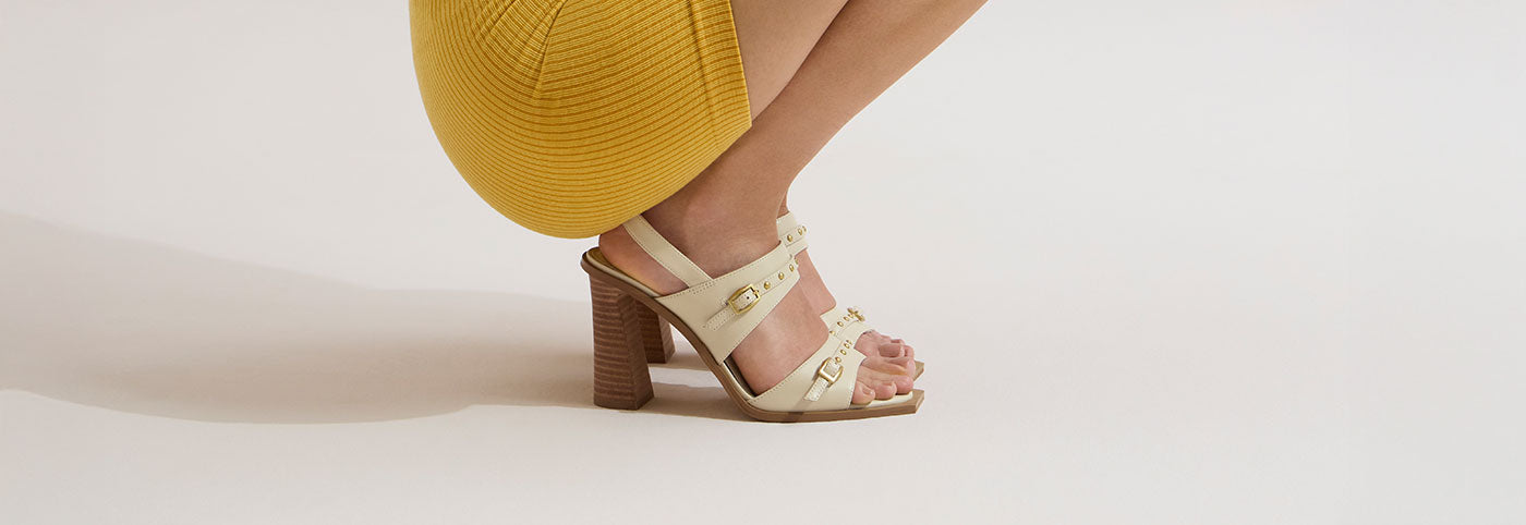 A person wearing a yellow ribbed skirt is crouched down, showing their legs and cream-colored high-heeled sandals with gold buckles on a light background.