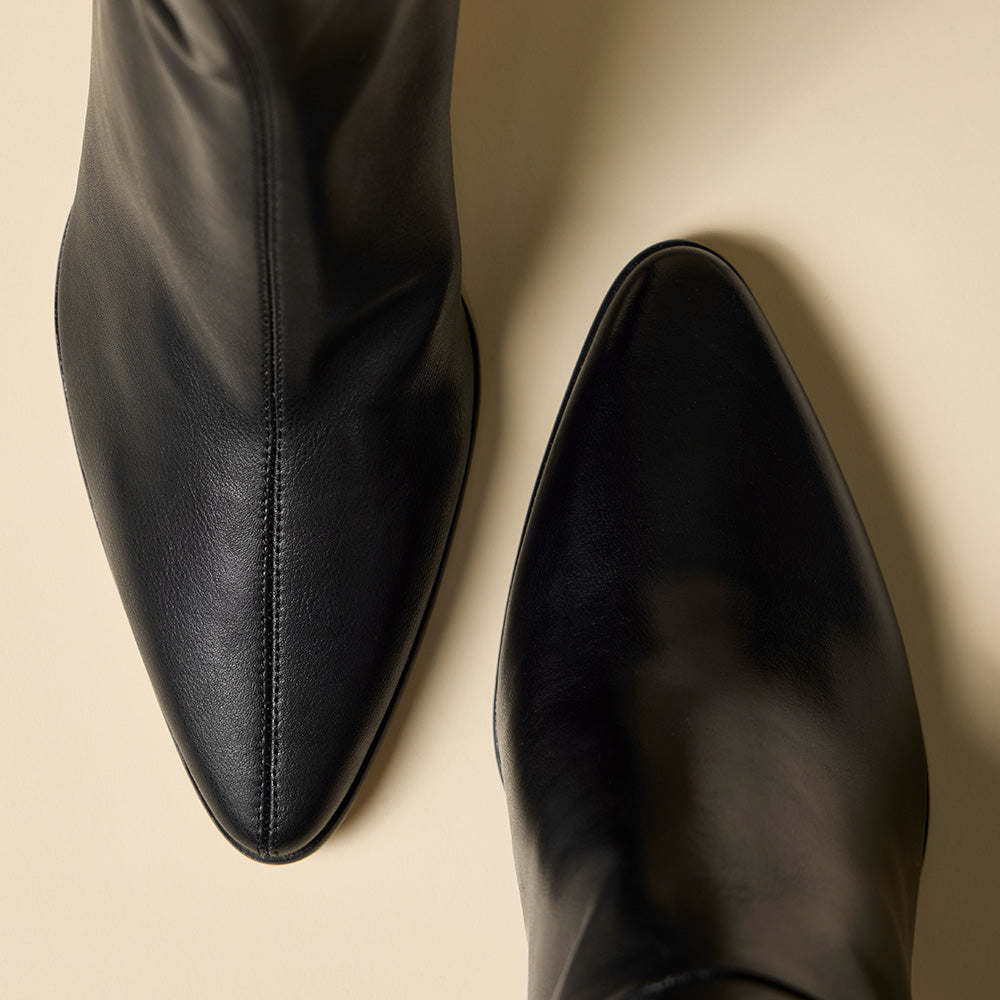 Close-up of a pair of black leather pointed-toe ankle boots on a warm beige background.