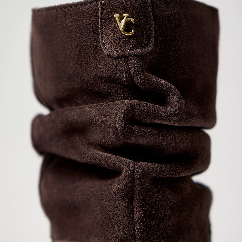 A close-up of a dark brown suede boot or sock-like textile with a small gold 'VC' logo patch near the top, shown against a light gray background.