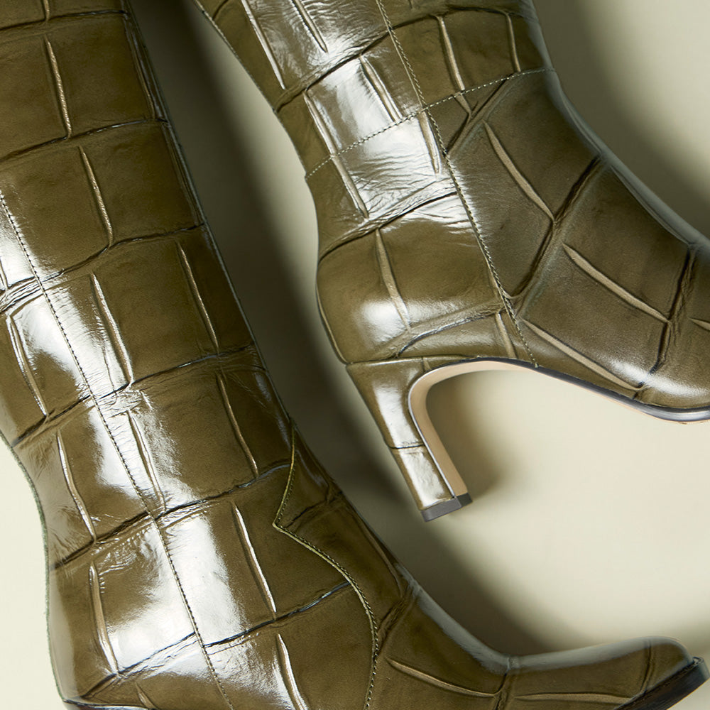 A pair of knee-high boots with a glossy, embossed crocodile or alligator pattern in a muted olive-brown color, shown against a neutral beige background.