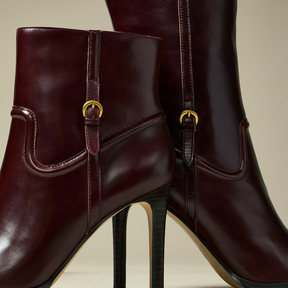 A pair of dark burgundy leather ankle boots with high stiletto heels and gold-tone buckle details, shown against a neutral beige background.