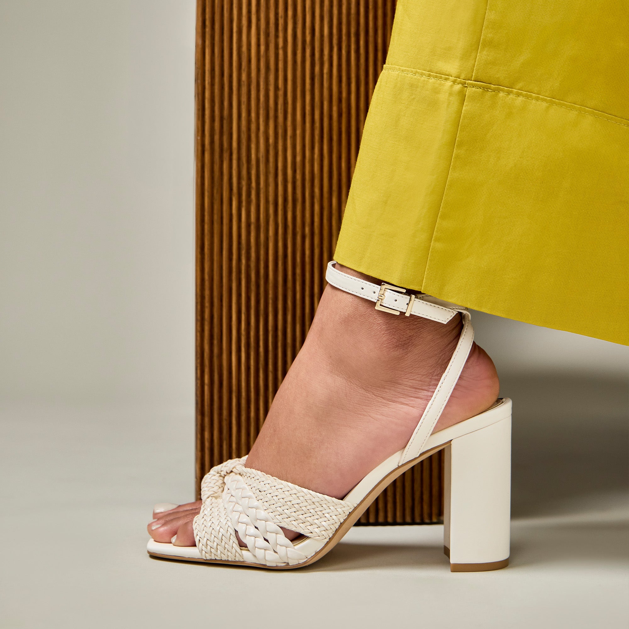 A close-up of a person's foot wearing a white woven slingback sandal with a block heel, paired with a yellow skirt, standing against a brown slatted panel.