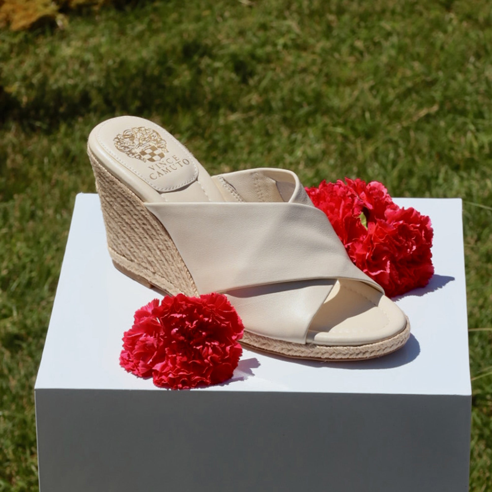 Pair of cream-colored wedge sandals on a white block with red flowers against a grassy background