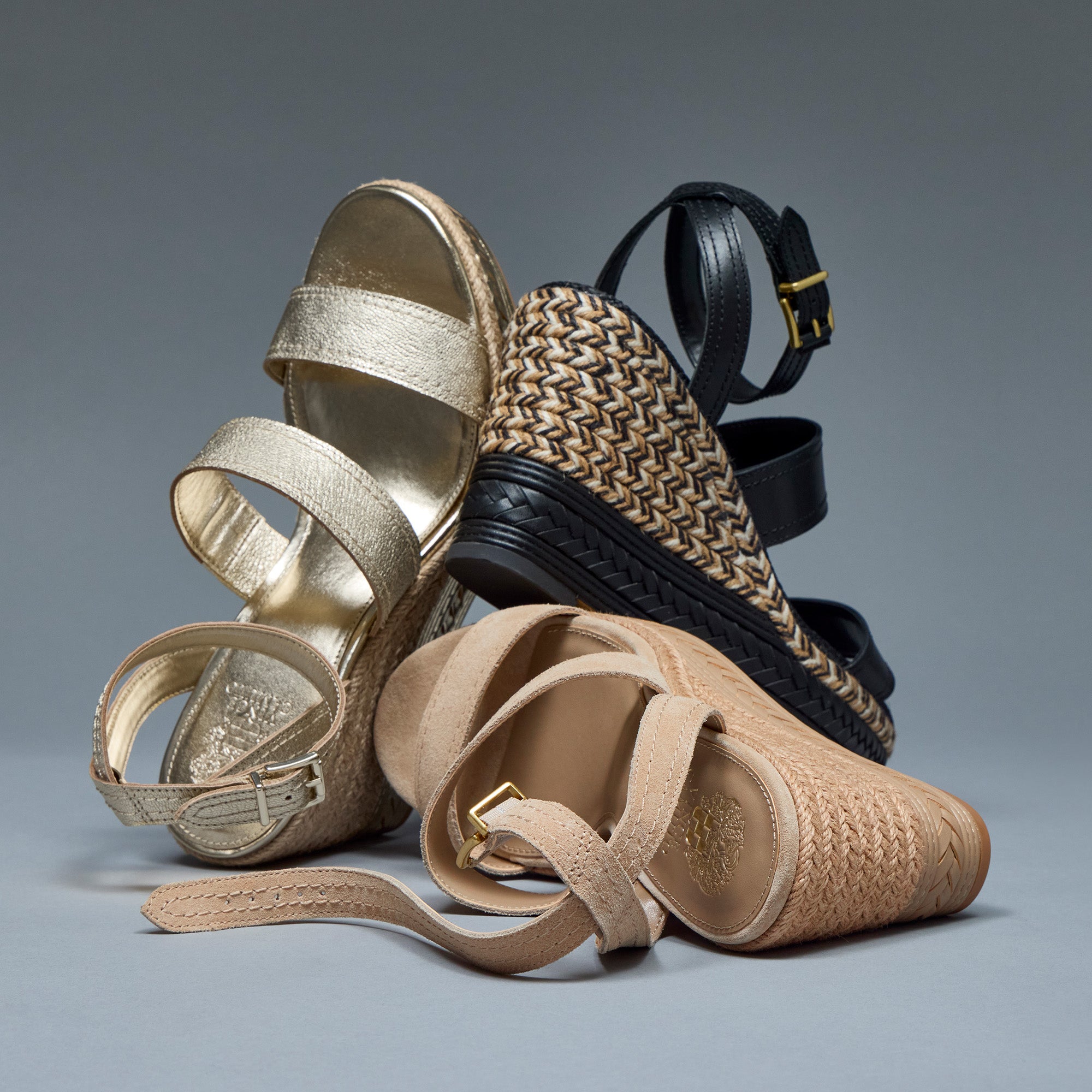 Three women's wedge sandals arranged on a smooth gray studio background: a metallic gold strappy sandal, a black ankle-strap wedge with woven beige detailing, and a beige woven espadrille-style sandal with an ankle strap.