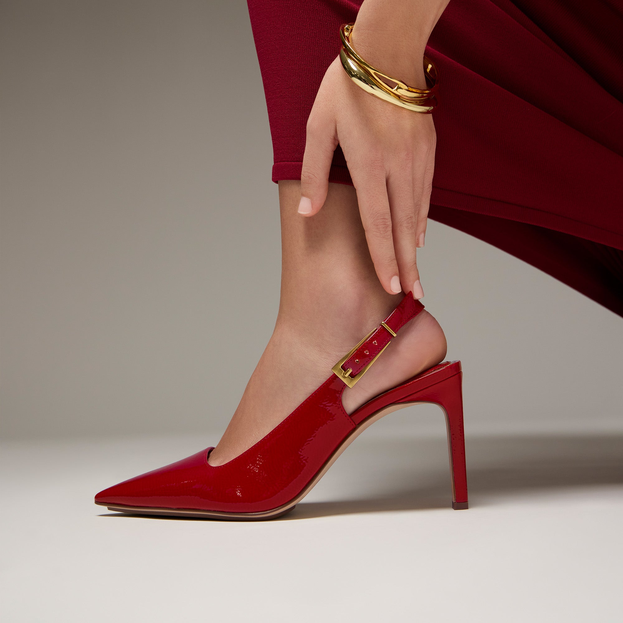 A close-up of a model's foot wearing a glossy red slingback high heel with a gold buckle, paired with a red dress and gold bangles, against a neutral studio background.