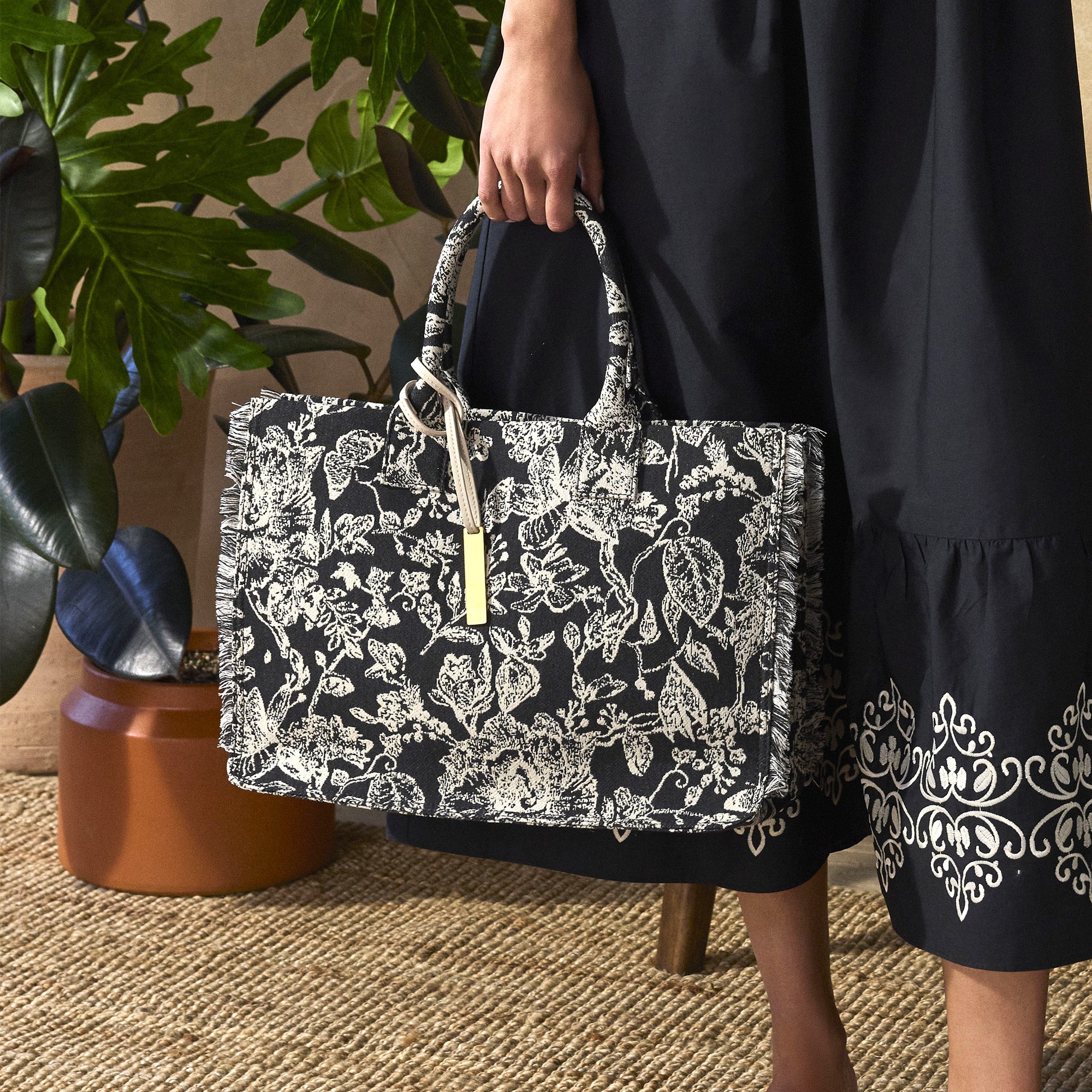Person holding a black floral-patterned handbag with a plant in the background