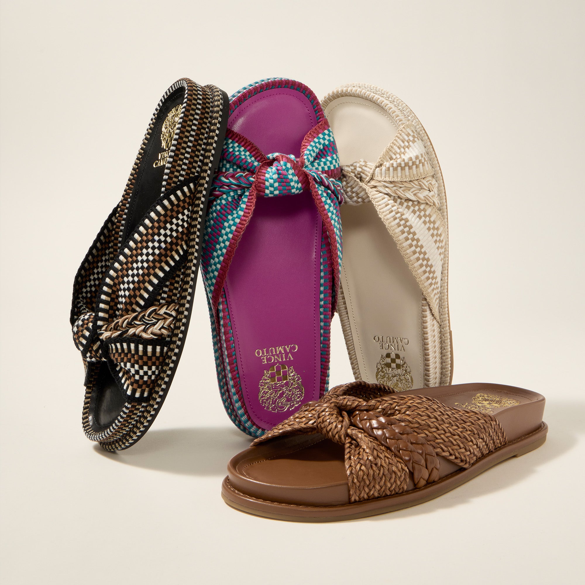 Four woven thong sandals arranged together on a light beige studio background. The sandals include black and white, purple and turquoise, off-white, and brown colorways, all with braided bow details and woven straps.