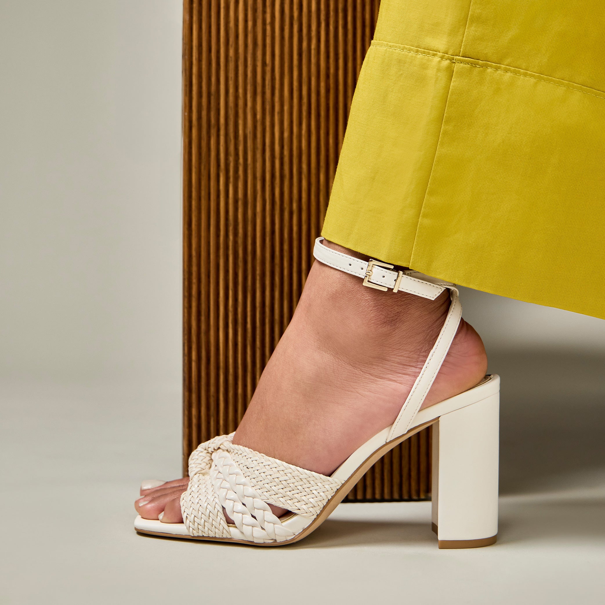 A person wearing a yellow pant and a white braided high-heeled sandal with an ankle strap stands next to a textured vertical surface.