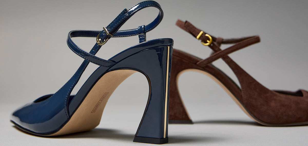 Two high-heeled mule sandals on a neutral gray studio background. The foreground sandal is glossy navy blue patent leather with a block heel and ankle strap.