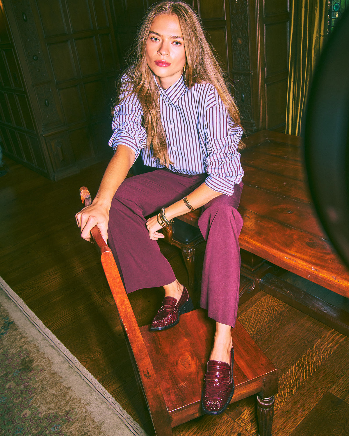 A woman sits on a wooden bench in a rustic room, wearing a blue and white striped button-down shirt, purple cropped trousers, and dark burgundy loafers.