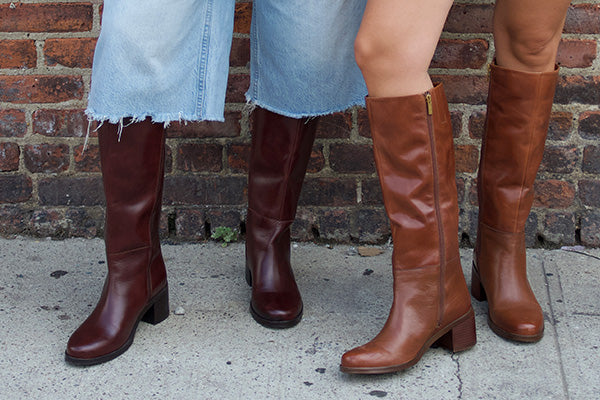 Three pairs of knee-high boots worn by models standing against a brick wall on a sidewalk. The boots include two dark burgundy boots and one medium brown boot, paired with light blue frayed-hem jeans.