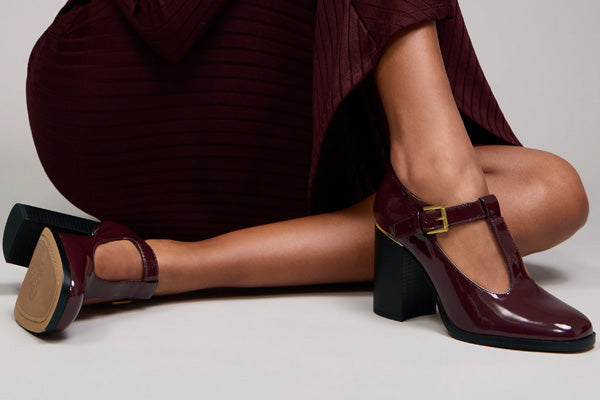 A pair of women's Mary Jane block-heel shoes in glossy burgundy patent leather with gold buckle details, shown on a model wearing a matching pleated burgundy dress against a light gray studio background.
