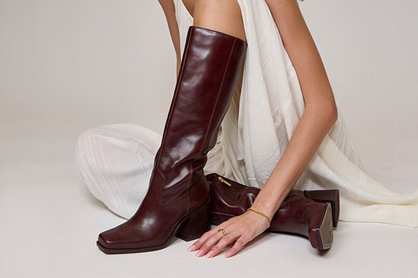 A model wearing a long, dark burgundy leather knee-high boot sits on a light beige studio floor, draped in a white dress. A second matching boot rests beside her hand.