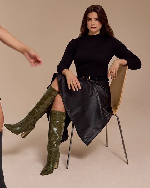 A woman in a black top and leather skirt sits on a chair, one arm draped over the back. She wears the Bennidy Knee High Boot by Vince Camuto in green, their block heel adding a modern edge to her ensemble. Another person’s arm and leg are partially visible on the left side.