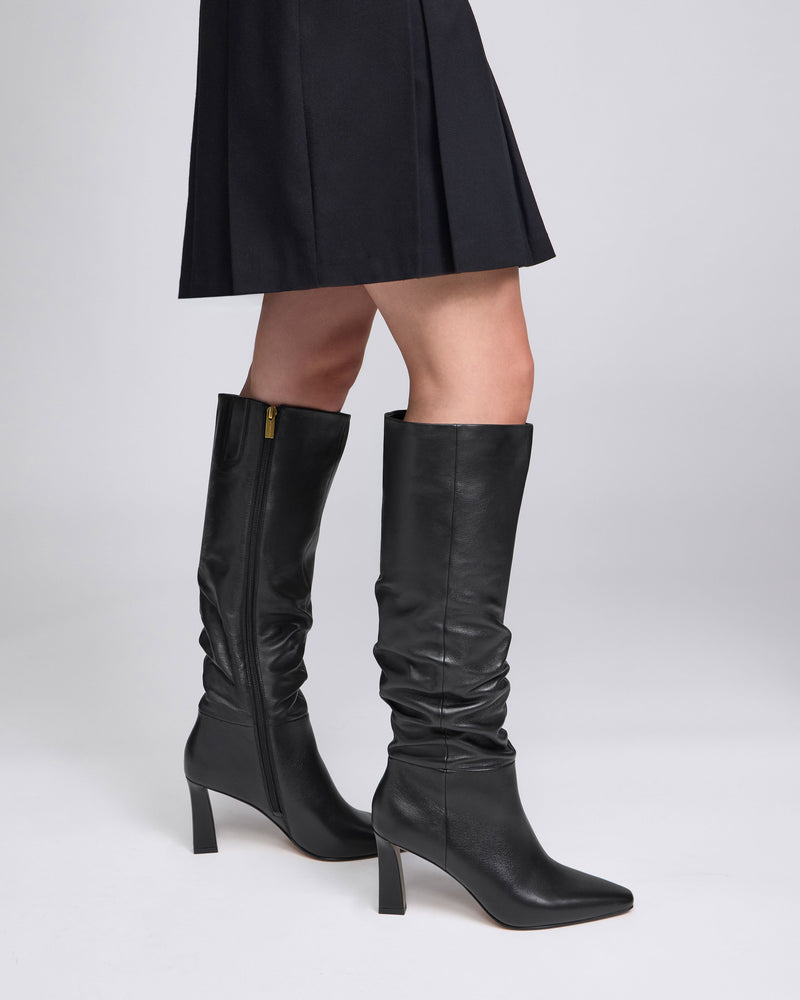 A person wearing a black skirt and the Vince Camuto Genisa Narrow Calf Slouchy Knee High Boots, standing against a plain light background.
