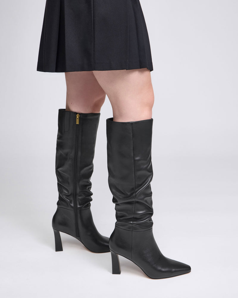 A woman wearing a black skirt and stylish Vince Camuto Genisa Slouchy Knee High Boots in black leather with a side zipper stands against a light gray background, with only the lower half of her body visible.