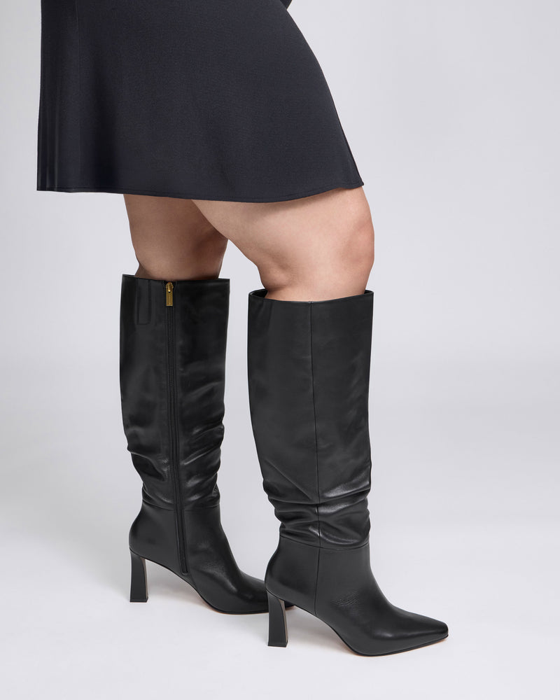 A person wearing a black skirt and stylish Vince Camuto Genisa Extra Wide Calf Slouchy Knee High Boots stands against a plain light background.