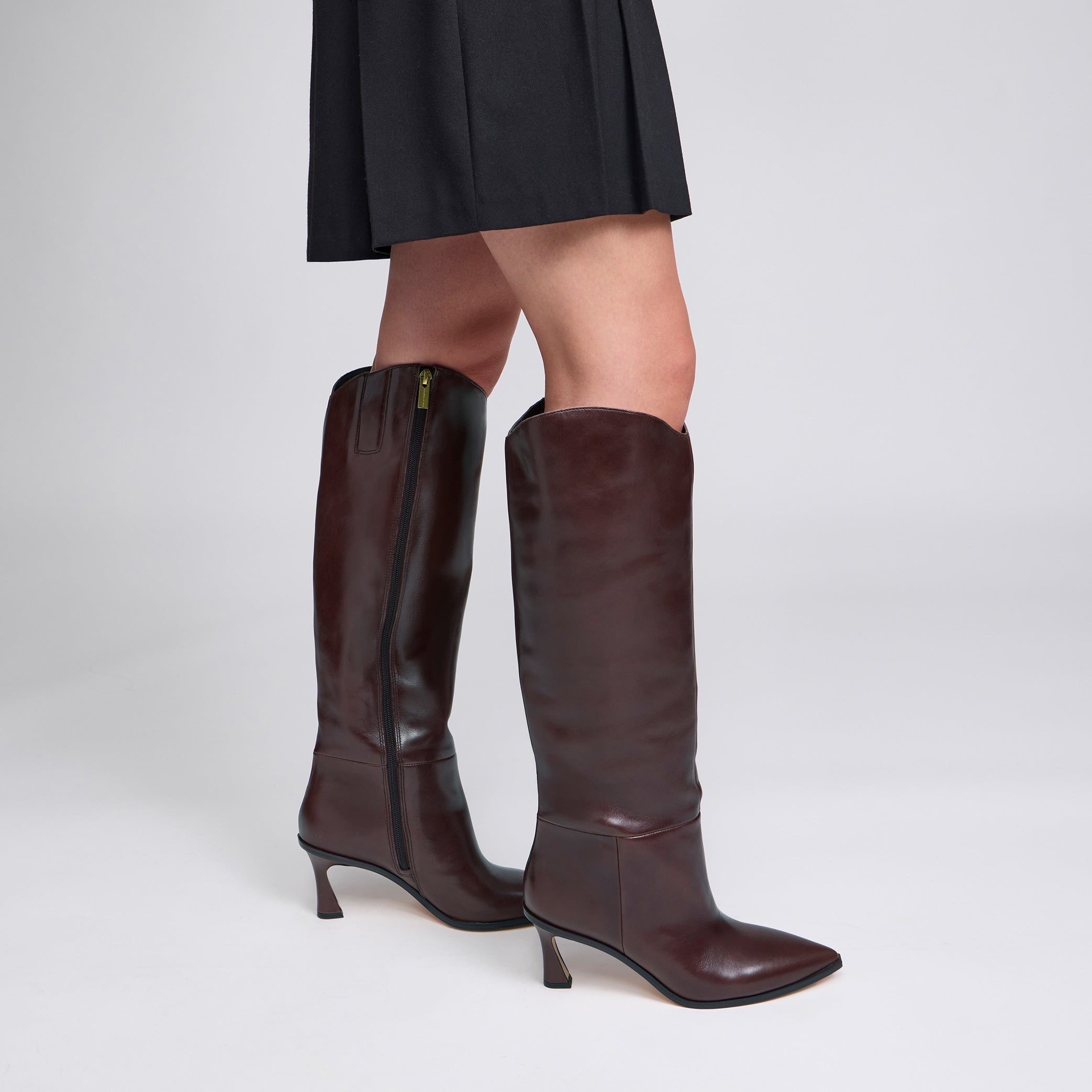 A pair of dark brown leather knee-high boots with pointed toes and kitten heels, shown on a model wearing a black pleated skirt against a light gray studio background.
