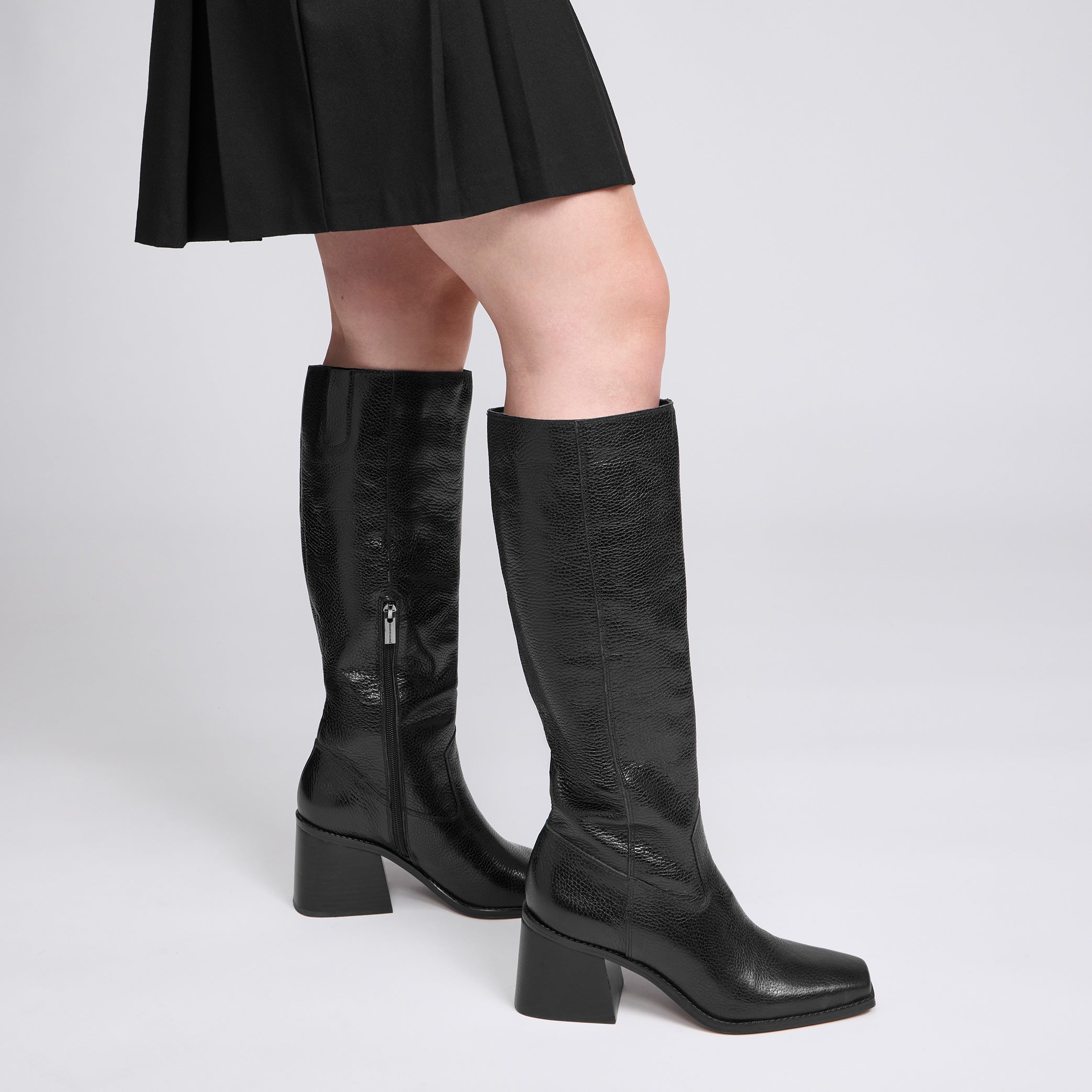 A pair of black knee-high leather boots with a block heel and side zipper, worn by a model in a black pleated skirt against a light gray studio background.