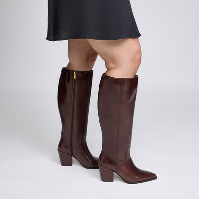 A pair of dark brown leather knee-high boots with a side zipper and block heel, shown on a model wearing a black skirt against a light gray studio background.