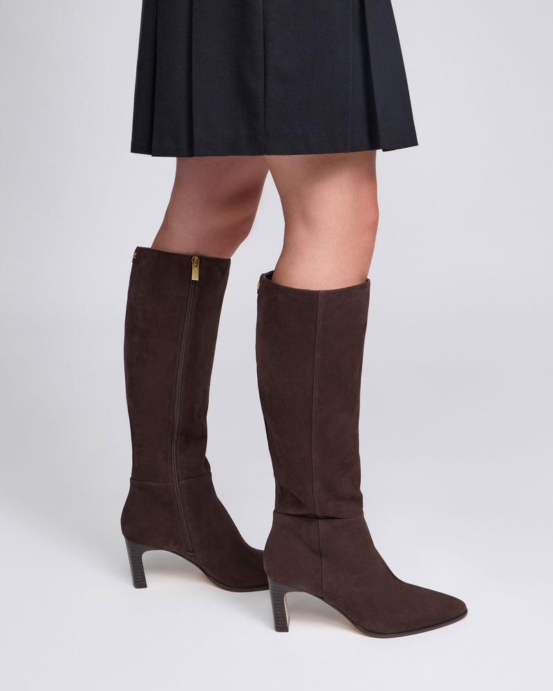 A person wearing the Vince Camuto Jorela Narrow Calf Knee High Boot in brown suede, featuring a gold zipper and block heel, paired with a black skirt, stands against a plain white background.