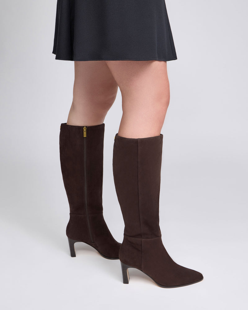 A person wearing a black skirt and the Vince Camuto Jorela Wide Calf Knee High Boot—featuring a block heel and a dark brown knee-high silhouette—stands on a light gray background.