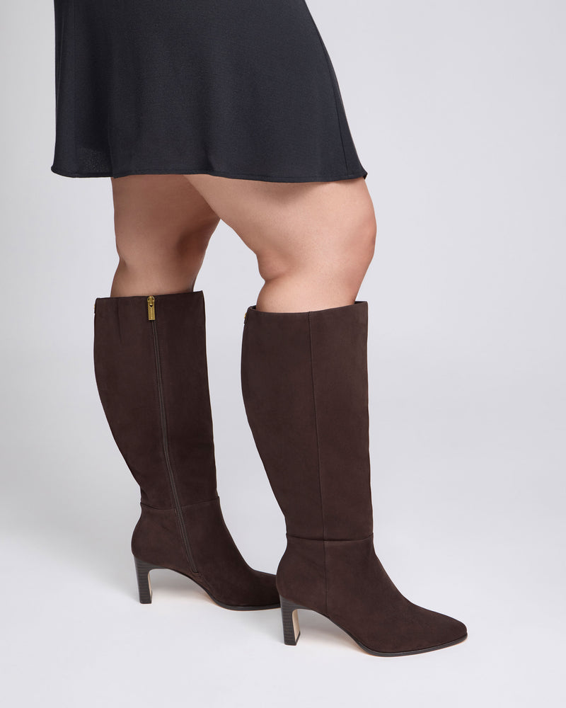 A person wearing a black skirt and the Vince Camuto Jorela Extra Wide Calf Knee High Boots, featuring a block heel and side zippers in dark brown, stands against a plain white background.