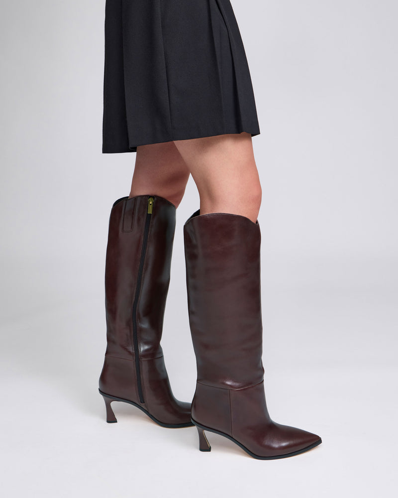 A person stands against a plain light background wearing a black skirt and the Vince Camuto Kenlea Narrow Calf Knee High Boot, which features dark brown leather, a side zipper, and low heels.
