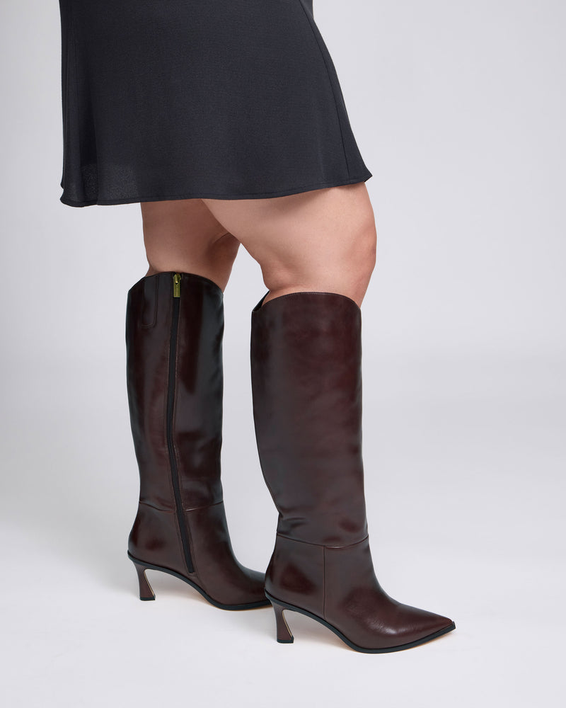 A person wearing a black skirt and Vince Camuto Kenlea Extra Wide Calf Knee High Boot in dark brown stands against a plain, light gray background. Only the lower half of the body is visible.