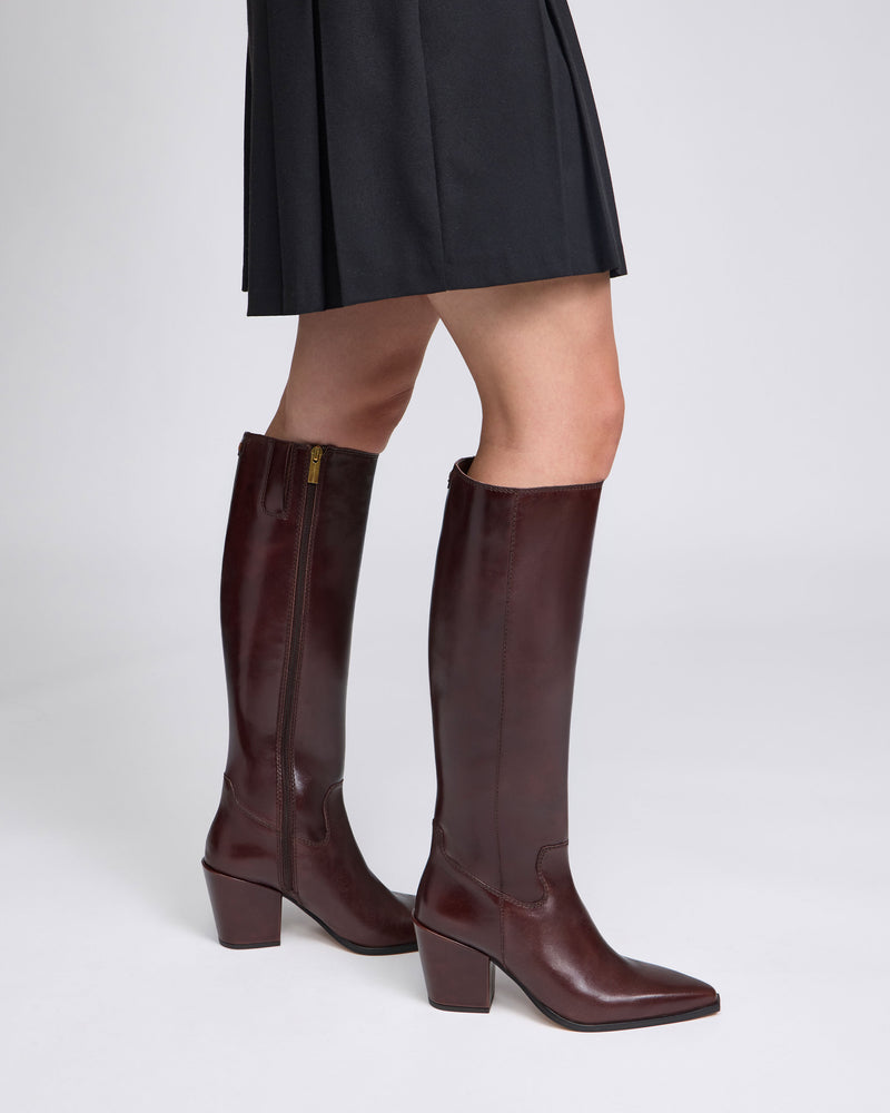 A person wearing a black skirt and the Vince Camuto Pamlia Narrow Calf Knee High Boots in dark brown leather with block heels stands against a plain light background.