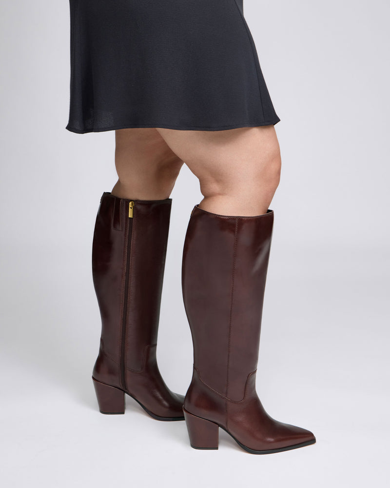 A person wearing a black skirt and the Vince Camuto Pamlia Extra Wide Calf Knee High Boot—featuring a stylish brown color, block heel, and pointed toe—stands on a light gray background.