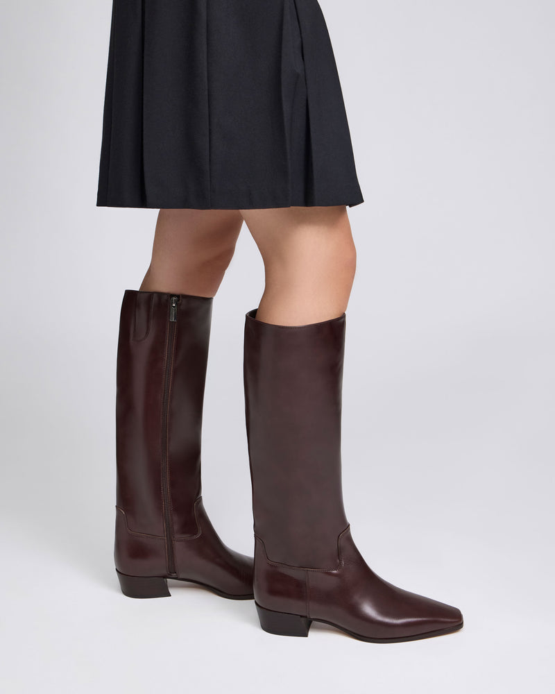 A person wearing a black skirt and the Vince Camuto Pavla Narrow Calf Knee High Boot—featuring a low block heel and subtle western styling—stands against a plain light background.