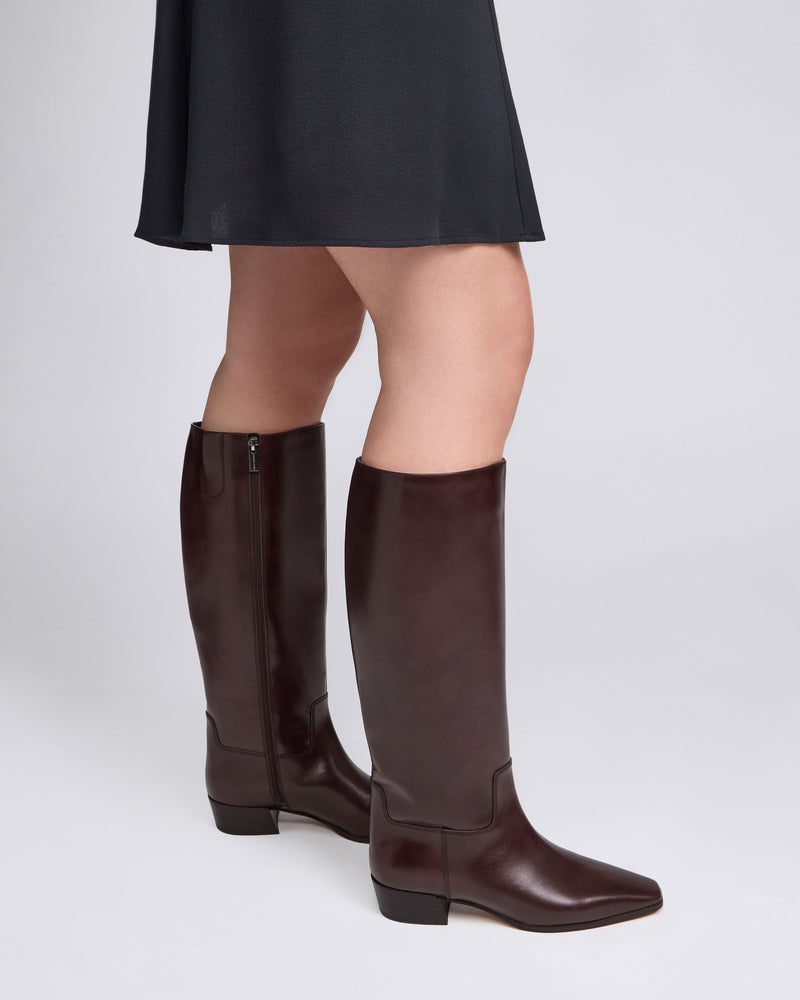 A person wearing a black skirt and the Vince Camuto Pavla Wide Calf Knee High Boots—tall, dark brown boots with low block heels and subtle western styling—stands against a plain light background.