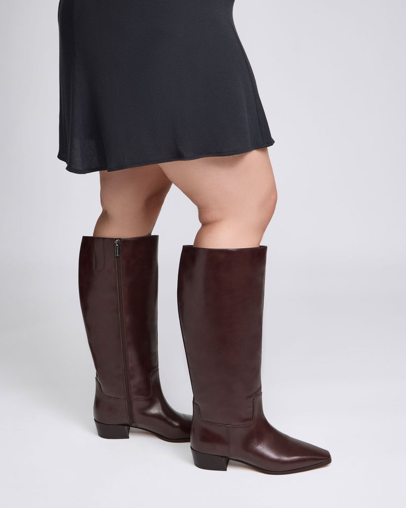 A person wears a black skirt paired with the Vince Camuto Pavla Extra Wide Calf Knee High Boot—knee-high, low-heeled boots in dark brown leather—standing against a simple, light background for a western-inspired look.
