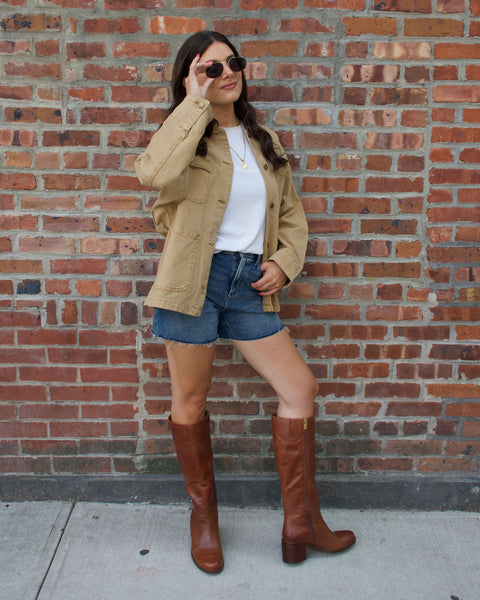 A woman wearing sunglasses, a tan jacket, white shirt, denim shorts, and the Vince Camuto Ammar Knee High Boot with a stylish block heel stands against a brick wall, posing with one hand on her sunglasses and the other in her pocket—a chic addition to any casual wardrobe.