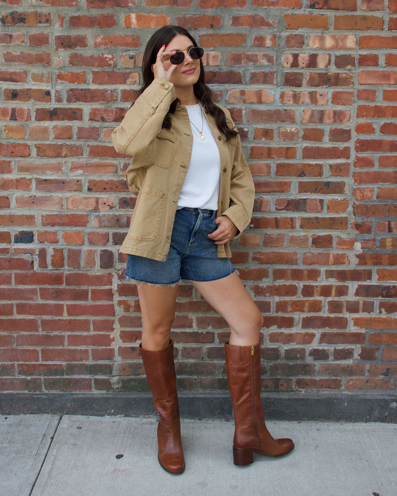 A woman poses against a brick wall, exuding effortless style in sunglasses, a tan jacket, white shirt, denim shorts, and the chic Ammar Narrow Calf Knee High Boot by Vince Camuto—her hand adjusting her sunglasses and the other on her hip—making these sophisticated brown boots with a block heel a standout addition to any casual wardrobe.