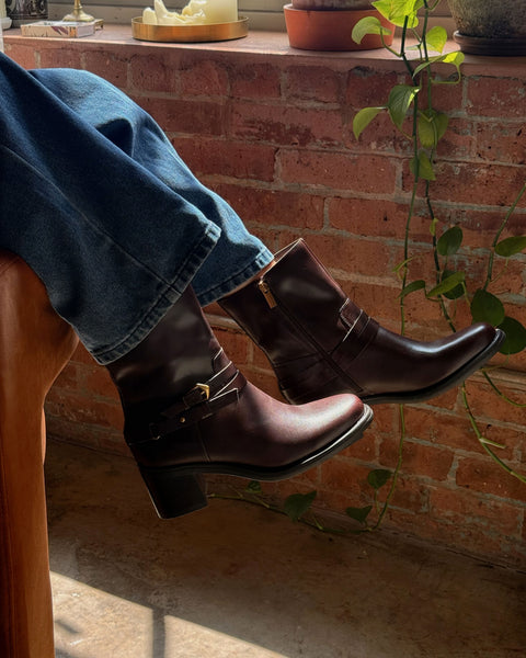 Wearing the Vince Camuto Armia Bootie in dark brown with blue jeans, a person rests their legs on a chair. Sunlight streams through a window, illuminating a backdrop of brick wall, potted plants, and a trailing vine.