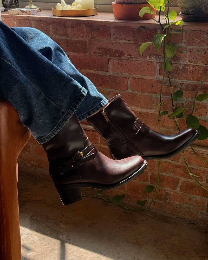 Wearing the Vince Camuto Armia Bootie in dark brown with blue jeans, a person rests their legs on a chair. Sunlight streams through a window, illuminating a backdrop of brick wall, potted plants, and a trailing vine.