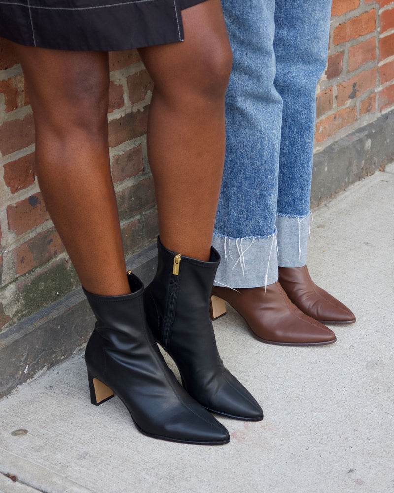 Two people stand side by side; one wears the Vince Camuto Julea Bootie—sleek black block heel ankle boots—with a skirt, while the other sports brown pointed-toe boots paired with blue cuffed jeans. They are standing on a sidewalk by a brick wall.