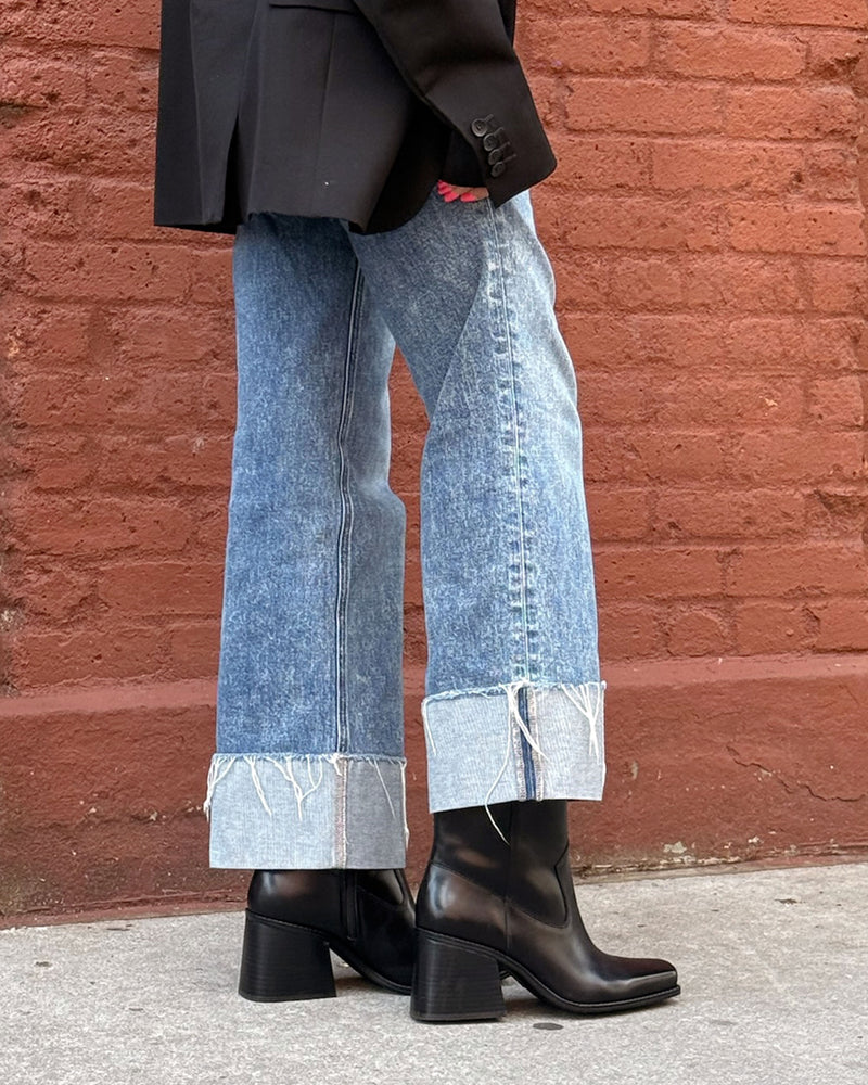 A person stands on a sidewalk wearing cuffed, frayed-hem jeans, a dark blazer, and the Vince Camuto Sybren Bootie—sleek black shoes with a chunky block heel. The red brick wall backdrop adds to the look’s modern sophistication.