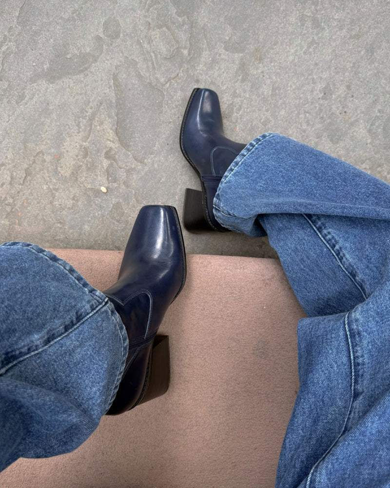 A person wearing blue jeans and the Vince Camuto Sybren Bootie—navy, western-inspired with a square toe and chunky block heel—sits with legs crossed on a light brown surface, gray floor underneath.