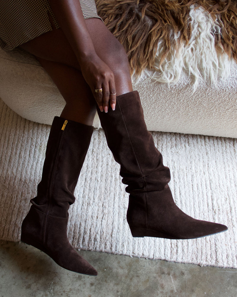 A person sits on a textured chair wearing the Vince Camuto Vanisa Extra Wide Calf Knee High Boots in brown suede, featuring pointed toes, a gold zipper, and a subtle wedge heel, with their feet resting on a cream-colored rug.