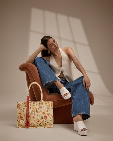 A woman sits on a rust-colored chair, wearing a sleeveless white top, wide-leg blue jeans, and white platform sandals. A floral tote bag with orange handles rests in front of her. Soft light casts window shadows on the wall.