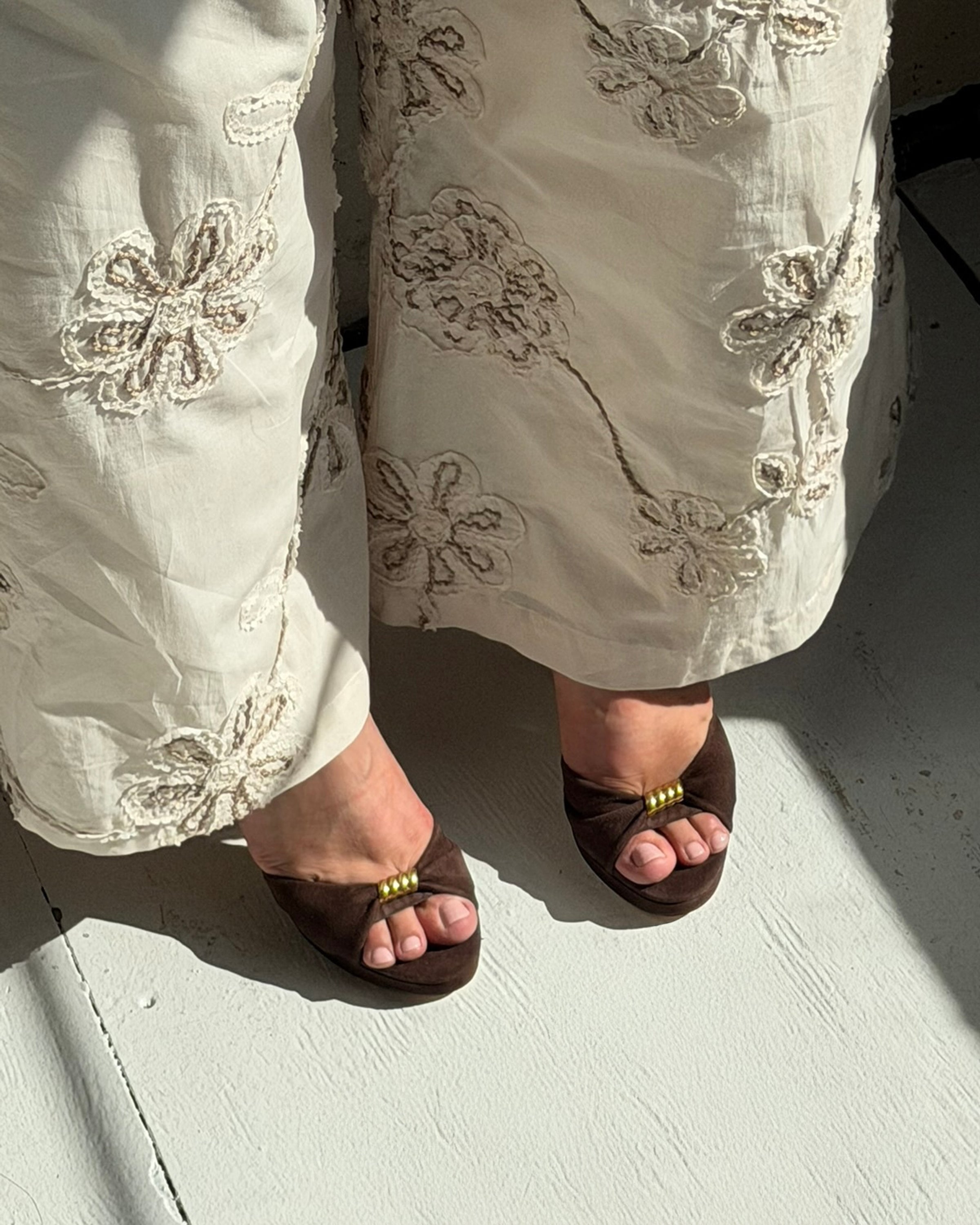 A person wearing cream-colored embroidered pants stands on a sunlit, light gray floor in the Neala Sandal by Vince Camuto.