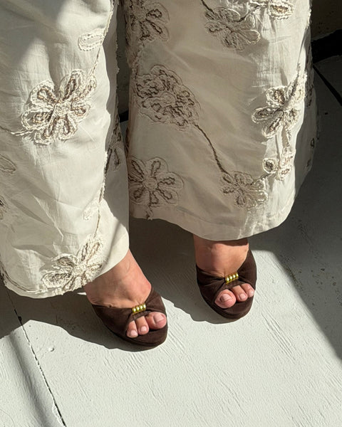 A person wearing cream-colored embroidered pants stands on a sunlit, light gray floor in the Neala Sandal by Vince Camuto.