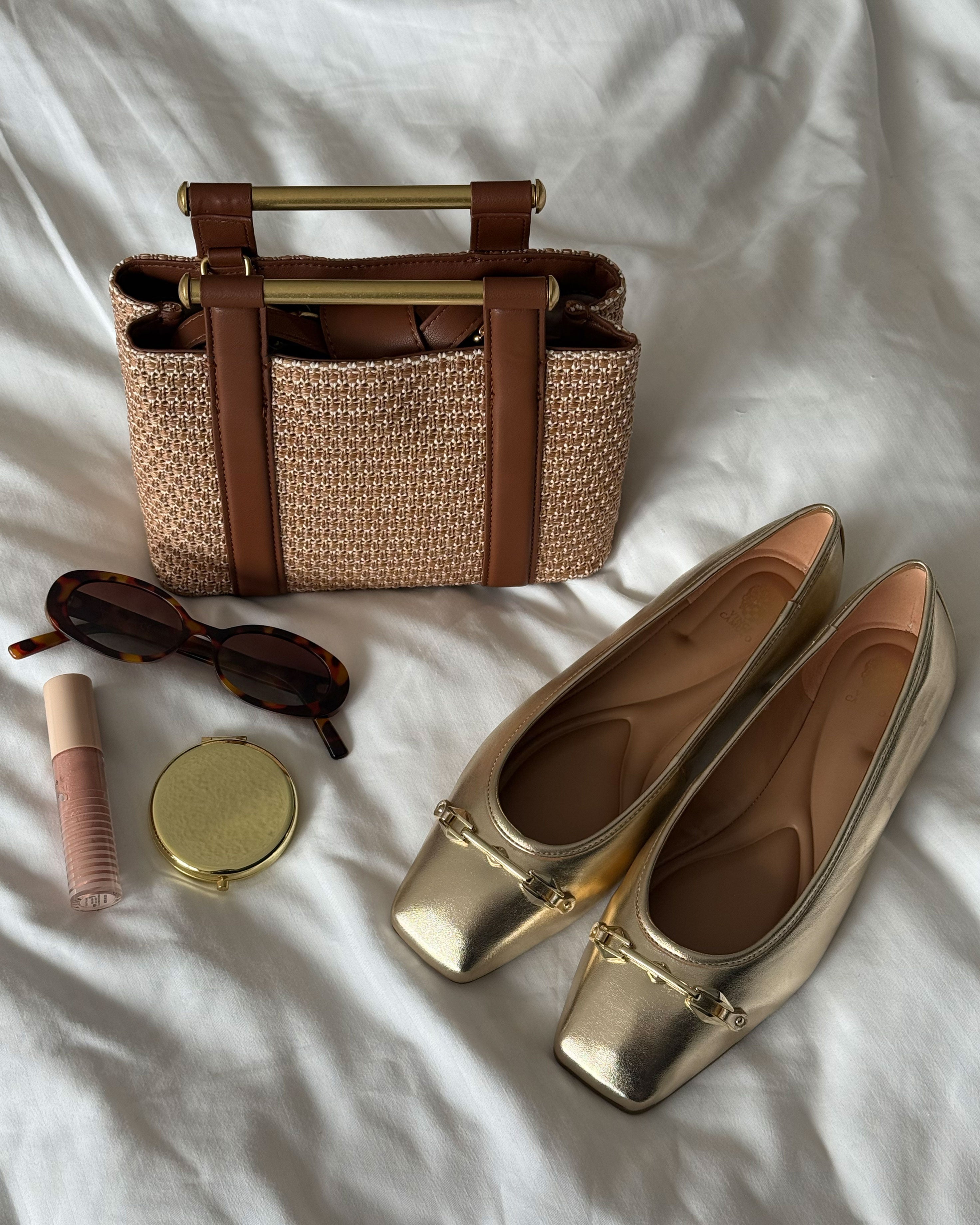 A small woven handbag with polished hardware and brown handles, tortoiseshell sunglasses, a gold compact mirror, nude lip gloss, and Vince Camuto's Virna Link Ballet Flat in metallic gold are arranged on a white fabric background.