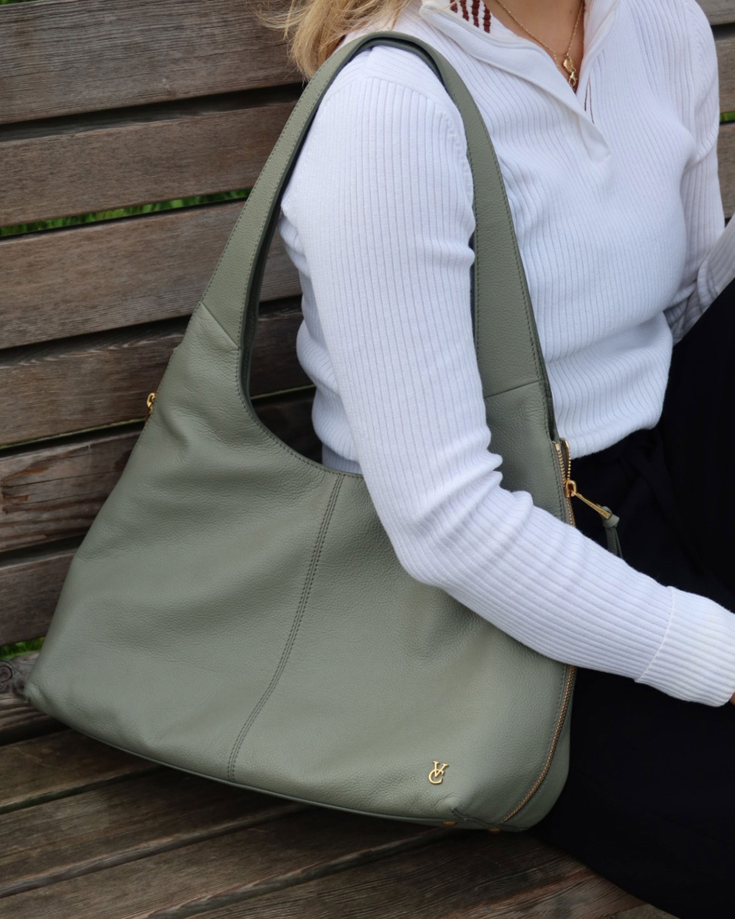 A person in a white ribbed top and black pants sits on a wooden bench, holding the Vince Camuto Kaela Tote Bag, a stylish sage green leather tote with gold zippers and a small gold emblem near the bottom corner.