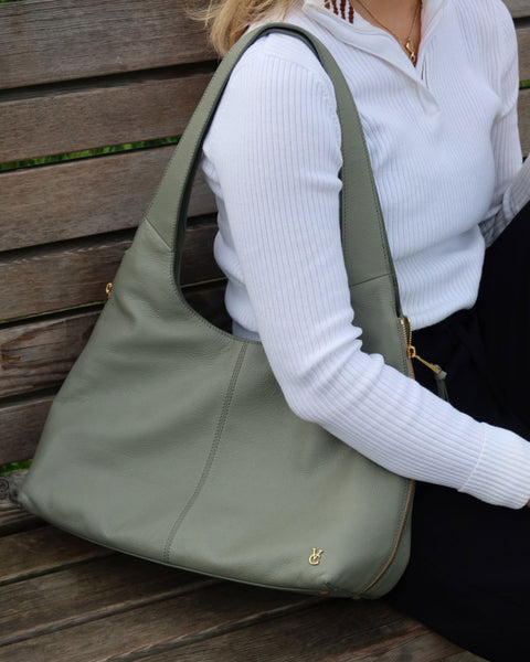 A person in a white ribbed top and black pants sits on a wooden bench, holding the Vince Camuto Kaela Tote Bag, a stylish sage green leather tote with gold zippers and a small gold emblem near the bottom corner.