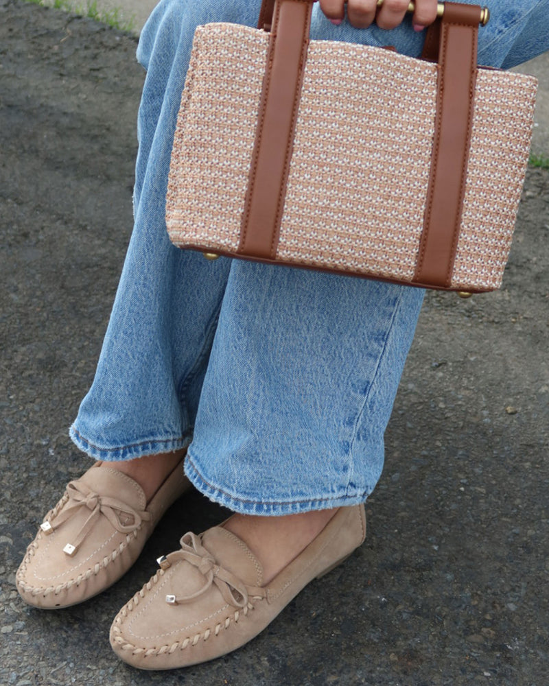 A person wearing light blue jeans and the Vince Camuto Katenas Flat Loafer in beige, featuring a moc-inspired silhouette, sits on the pavement holding a beige woven handbag with brown leather straps.